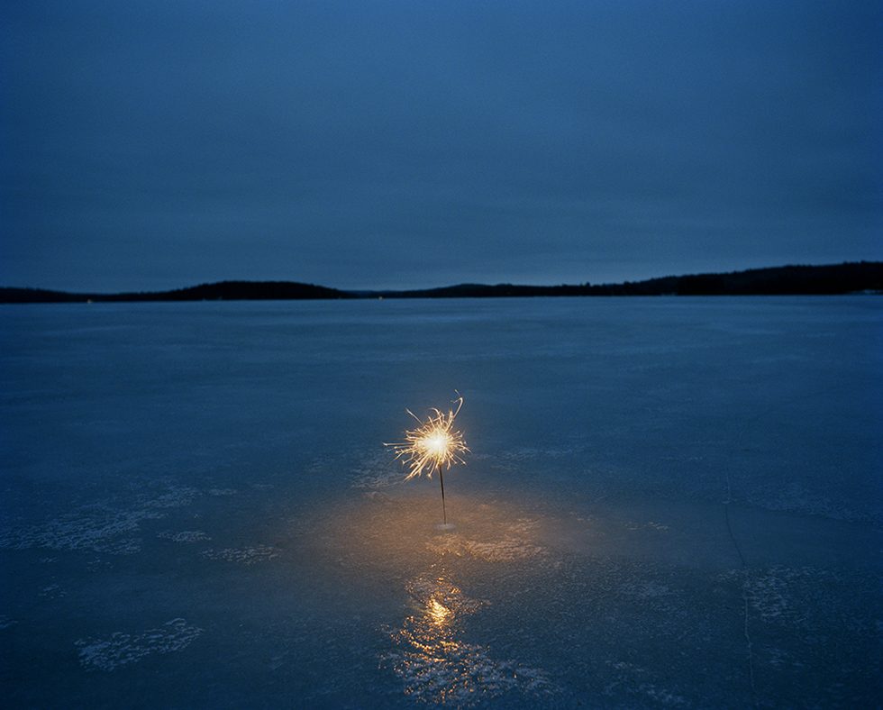 This is a color photograph of a burning sparkler stuck into a sheet of ice in a landscape setting with mountains in the distance.