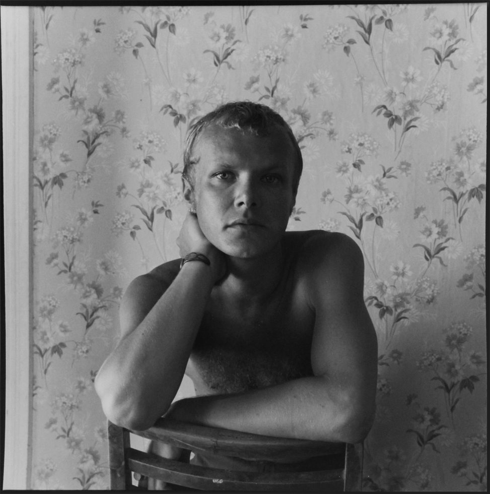 This is a black-and-white photographic portrait of a blond man sitting facing the back of a chair with his hand resting on his neck.