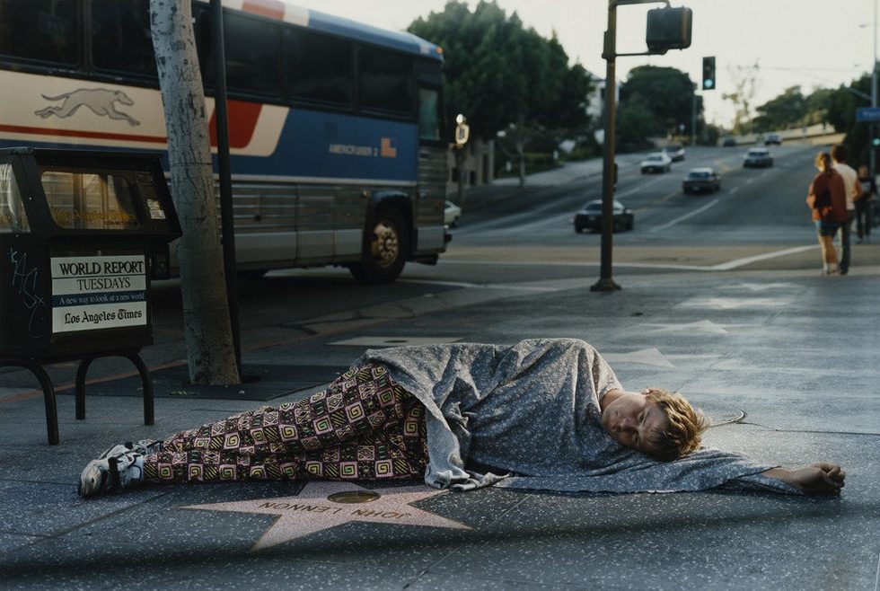 A color photograph of a homeless man sleeping next to John Lennon's star on the Walk of Fame in Los Angeles.