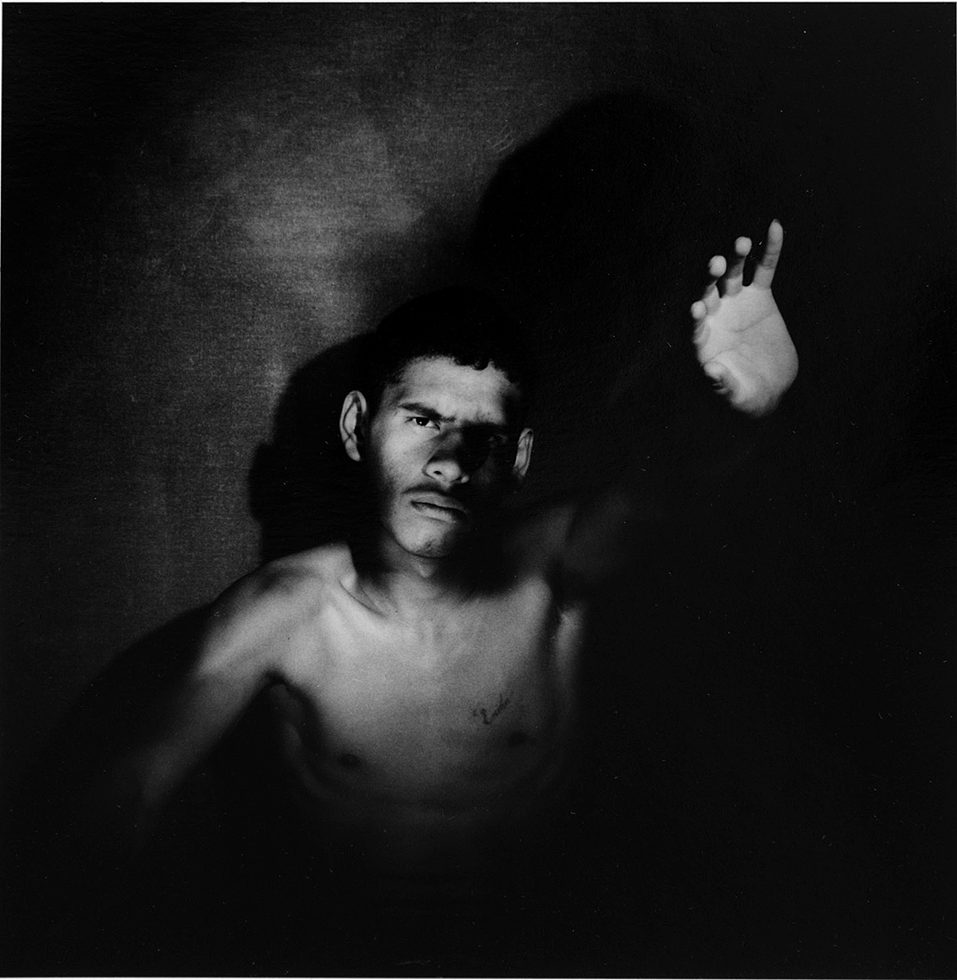 This is a black-and-white photograph of a man with a mustache in a dark space with his left hand raised.