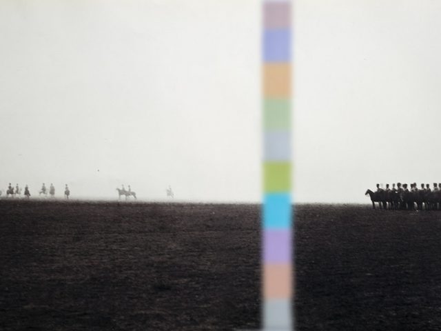 A black and white photograph of men on horseback in a field with a multicolored stripe going down the picture plane.