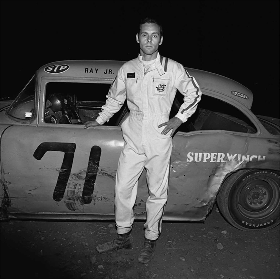 This is a black-and-white photograph of a man in a racing suit in front of a car with the number 71 painted on the door.