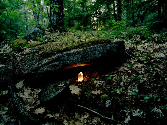 This is a color photograph of a forest floor with a glowing crystal beneath the ledge of a rock.