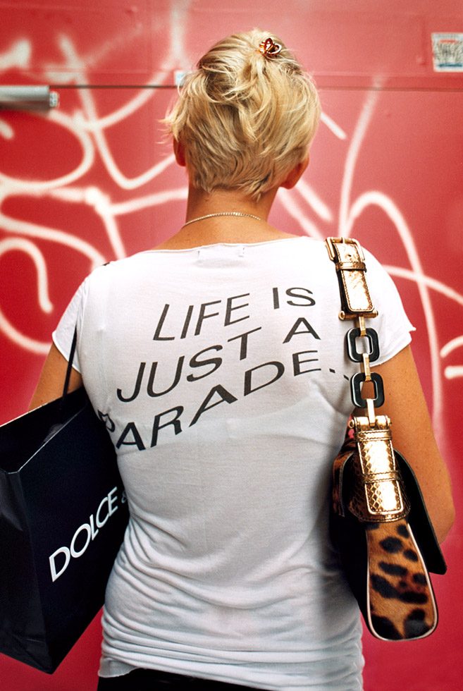 This is a color photograph of a blonde woman from behind wearing a graphic t-shirt with text against a red graffiti wall.