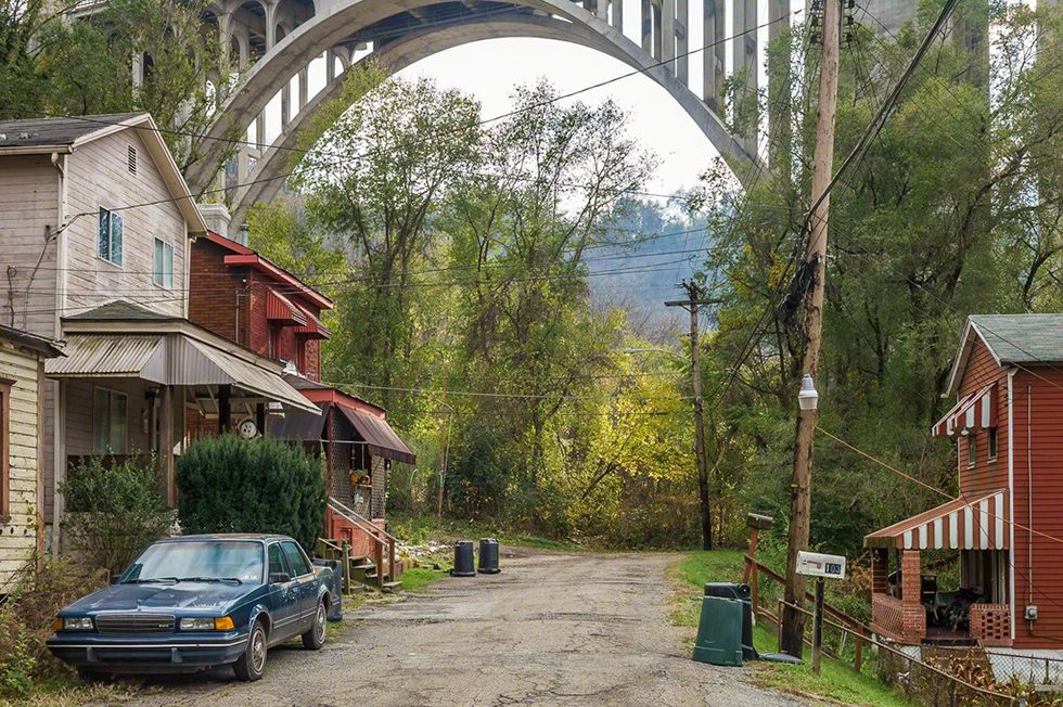 This is a color photograph of old houses and a blue car beneath a large bridge in the summer.