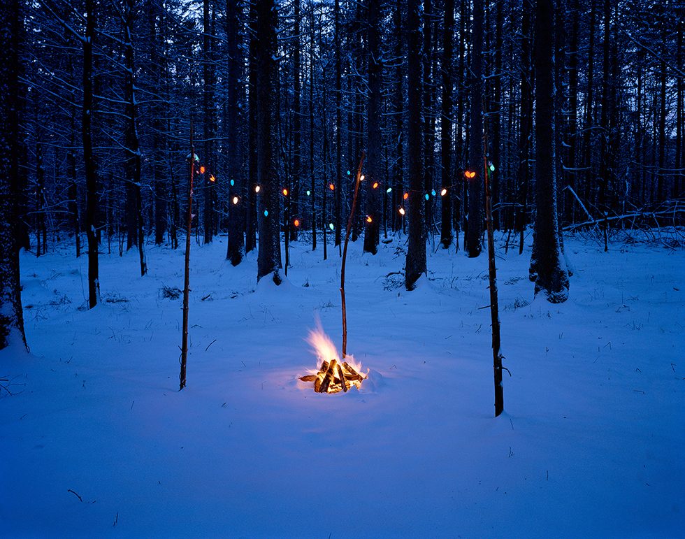 This is a color photograph of a campfire in a snowy landscape at dusk with strands of Christmas lights strewn from three poles above the fire.