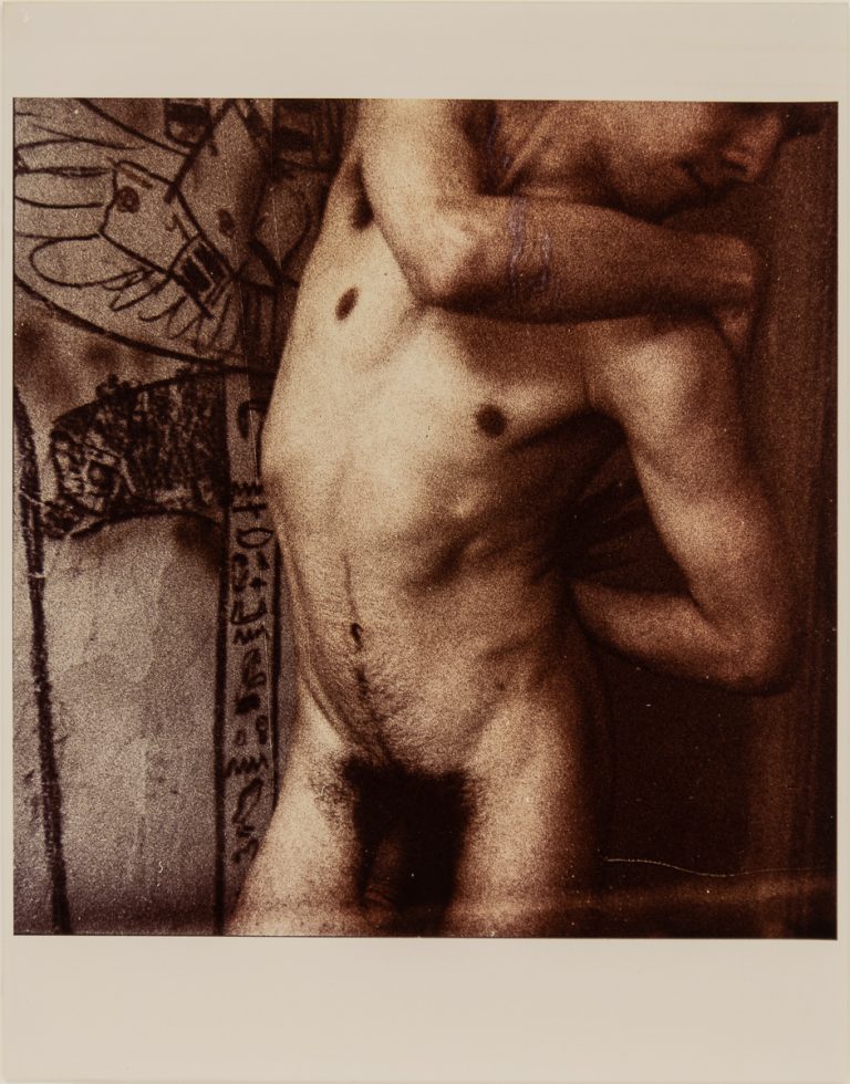 Sepia-toned photo of a nude figure beside a wall with abstract drawings and symbols.