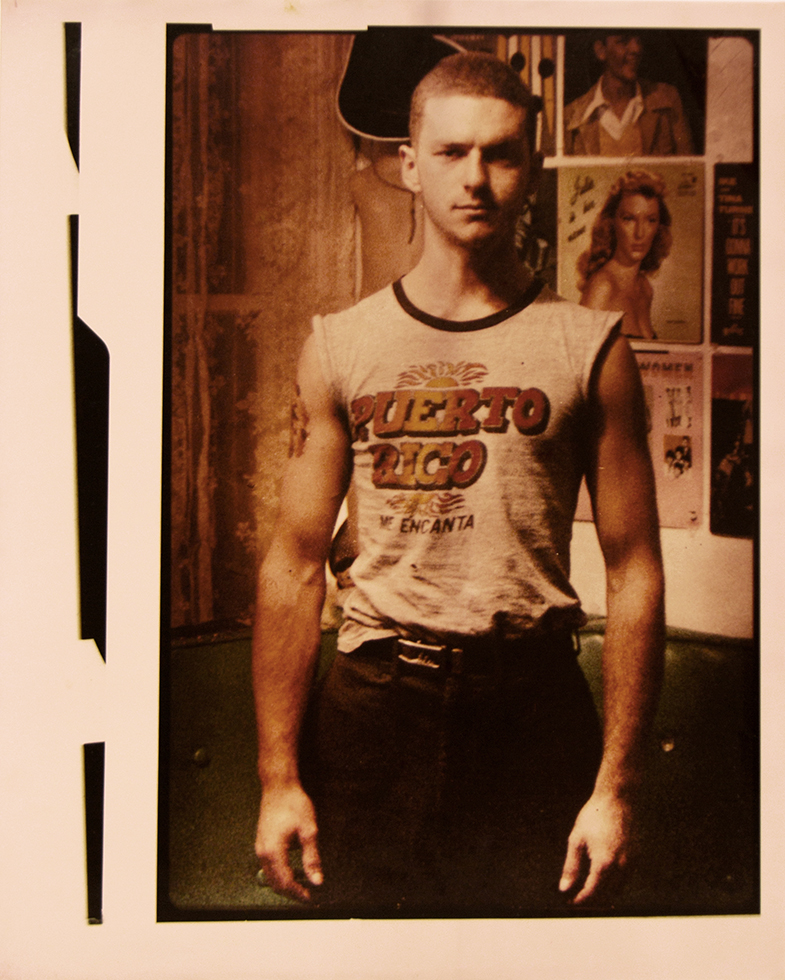 This is a muted color photograph of a man in a sleeveless Puerto Rico t-shirt standing in front of a wall with vinyl album covers.