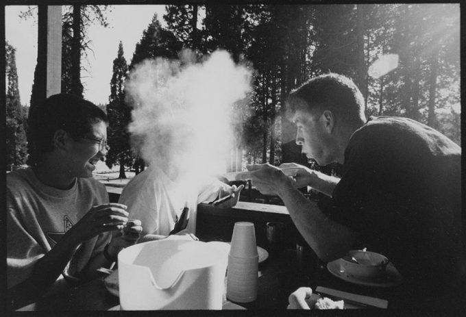 This is a black-and-white photograph of three teenage boys outside by pine trees blowing dust to make smoke-like clouds.