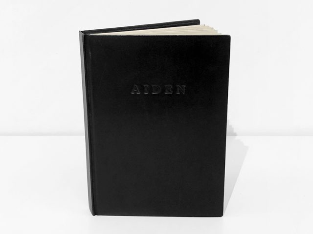 This is a photograph of a black-leather bound book against a white background that reads "Aiden" embossed on the cover. again