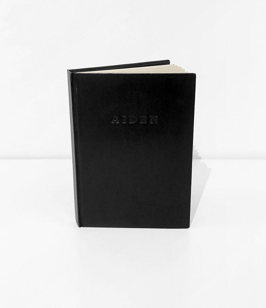 This is a photograph of a black-leather bound book against a white background that reads "Aiden" embossed on the cover. again