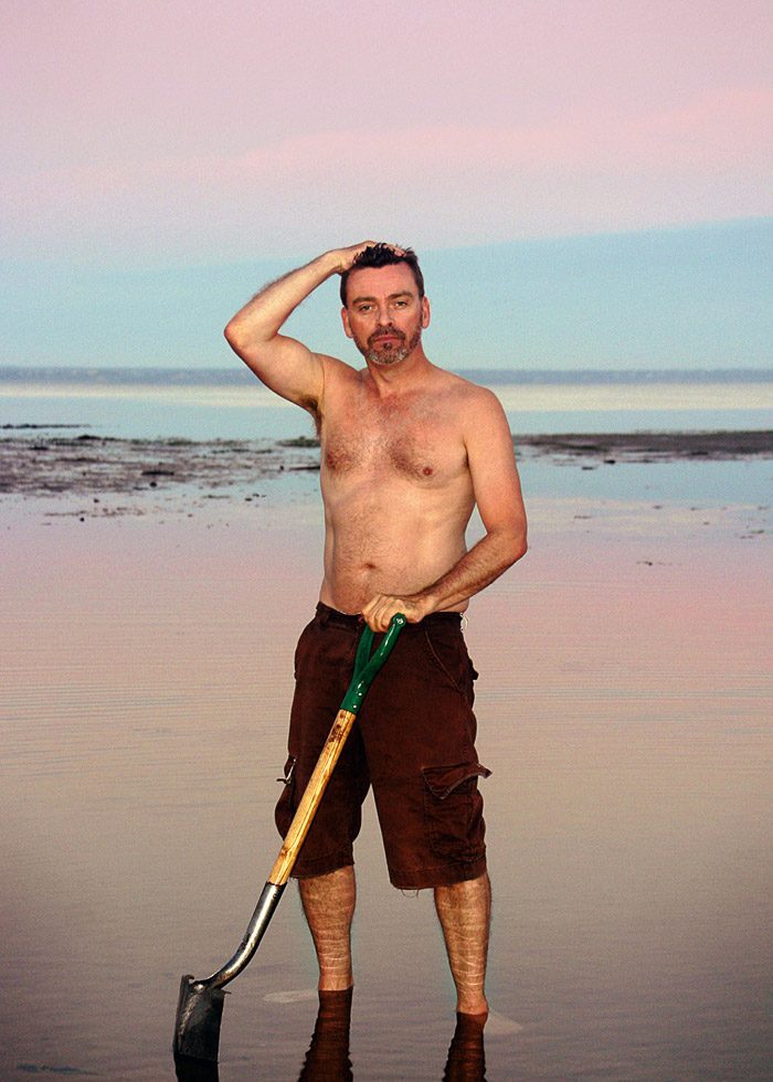 Shirtless self portrait by Frank Yamrus on a beach of ankle-deep water digging clams with a pink and blue sunset in the background.