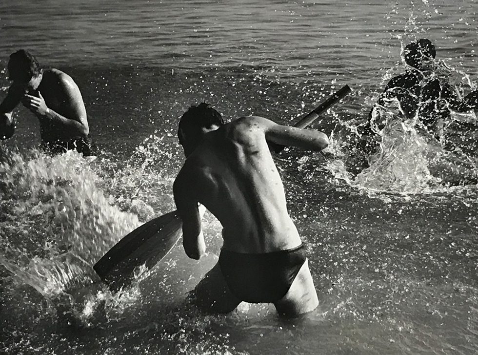 This is a black-and-white photograph of a man from behind in the sea splashing two other men facing the camera with an oar.