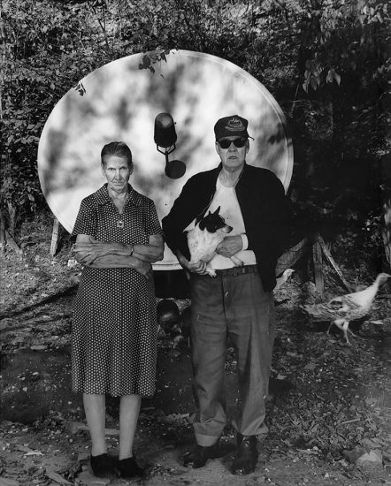 This is a black-and-white photograph of an old couple in front of a satellite dish.