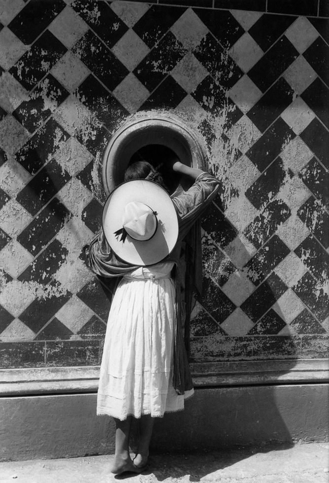 This is a black-and-white photograph of a girl in a white dress seen from behind while she is peeking through a dark circular window.