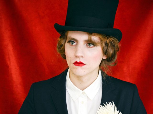 An image of a person in a black coat and top hat with a flower in their label against a red fabric backdrop.