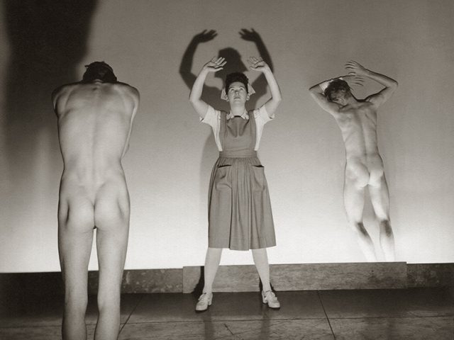Two nudes with their backs to the camera and a person wearing an apron facing the camera with their arms reaching towards the ceiling