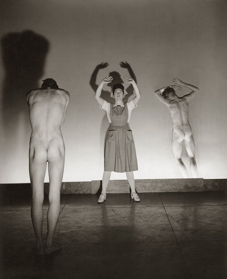 This is a black-and-white photograph of two male nudes with their backs to the camera and a woman wearing an apron and nurse cap facing the camera with her arms reaching towards the ceiling.