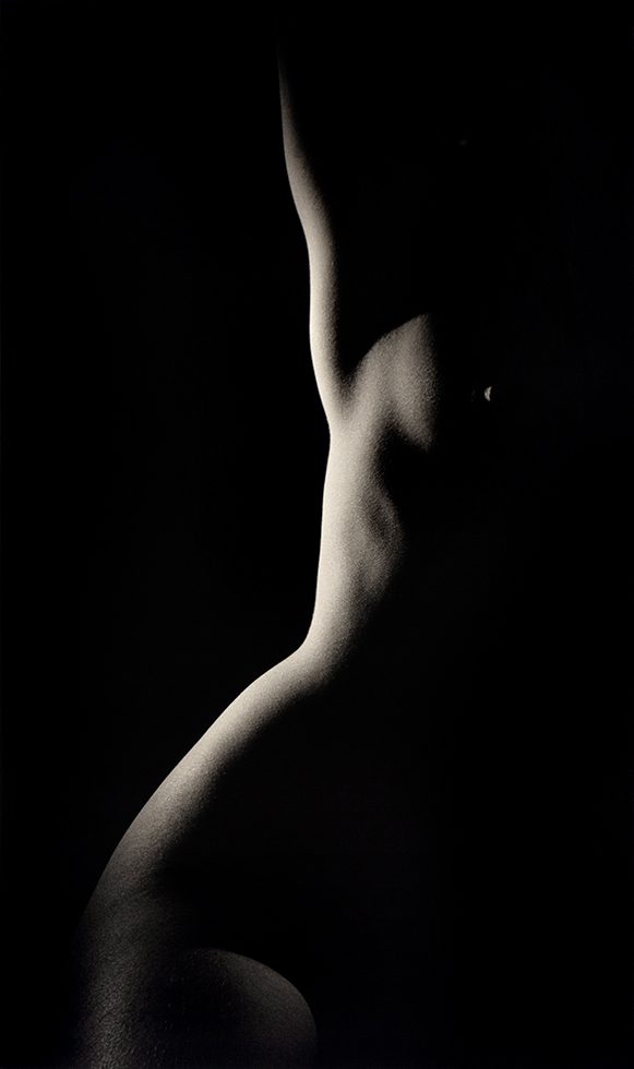 This is a warm-toned black-and-white photograph of one half of a naked woman's dramatically illuminated torso with her arm raised.