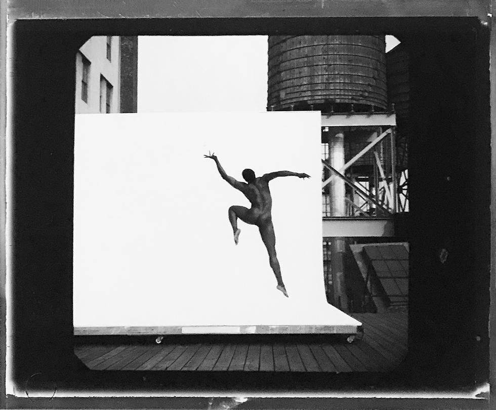 This is a black-and-white photograph of a nude Black dancer from behind leaping through the air on a New York rooftop against a white backdrop.