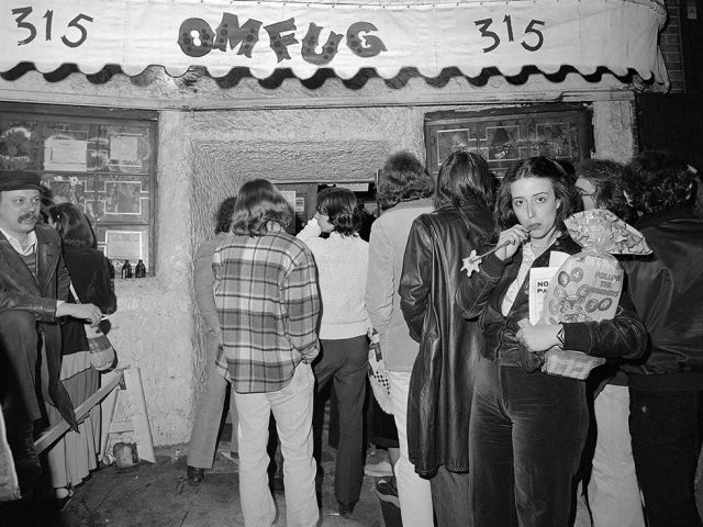 This is a black-and-white photograph of a woman sucking on the stem of a flower on the sidewalk outside of a dance club in New York City.