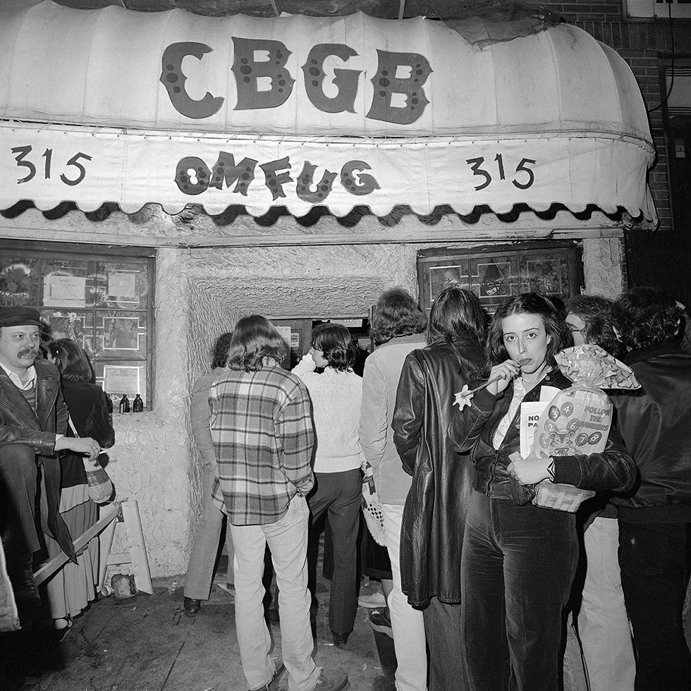 This is a black-and-white photograph of a woman sucking on the stem of a flower on the sidewalk outside of a dance club in New York City.