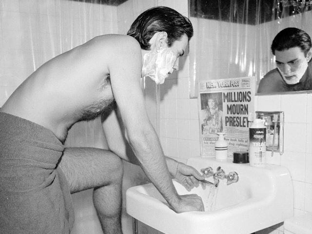 This is a photograph of a man shaving in a bathroom mirror with a newspaper.