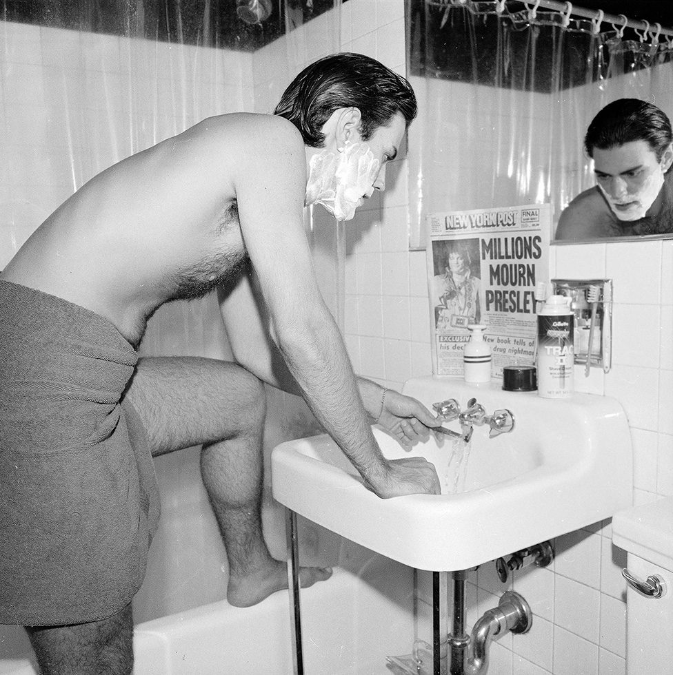 This is a photograph of a man shaving in a bathroom mirror with a newspaper.