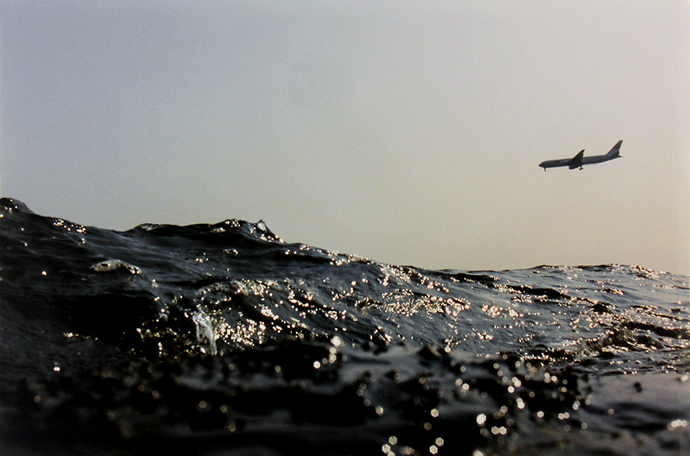 This is a color photograph of a plane in the sky with a big wave of water dominating the foreground of the image.