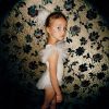 This is a color photograph of a young girl in profile wearing a white dress against a floral wall.