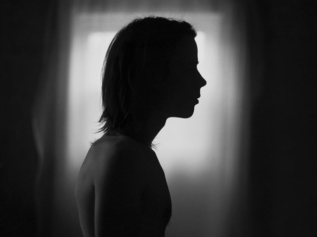 This is a black-and-white photograph of a nude man in profile in a dark room against a window with bright light behind him.