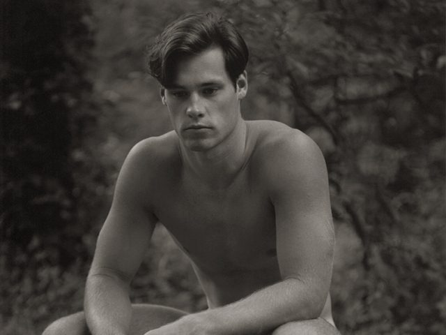 Black and white photograph of a nude young man crouching in the grass