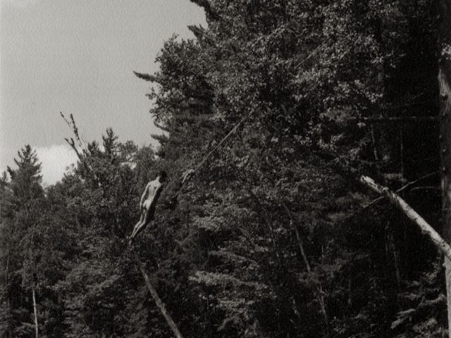 Black and white photograph of a nude man swinging on a rope swing above a lake about to fall into the water