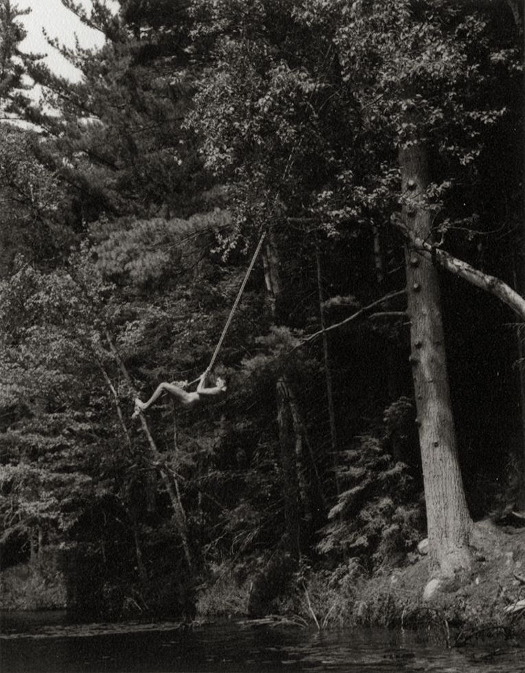 Black and white photograph of a man swinging on a rope above a lake