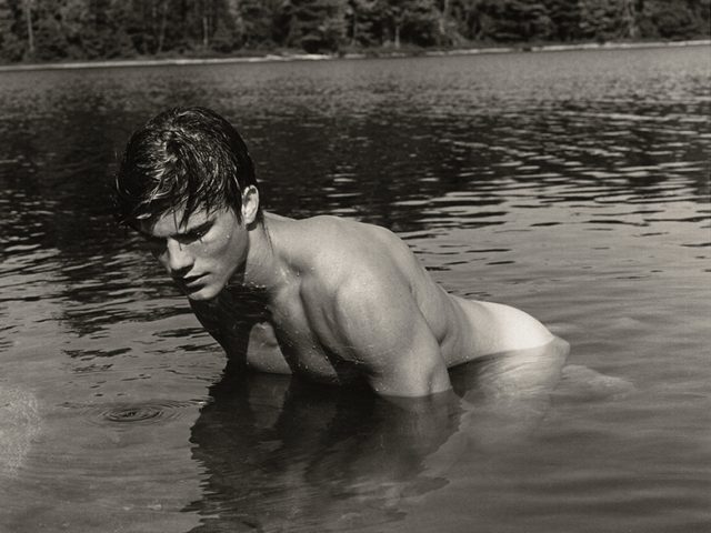 Black and white photograph of a man on all fours in a lake looking down at the water