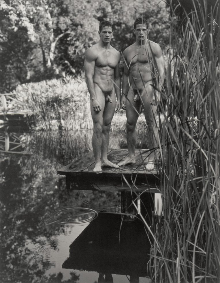 A vertical photograph by Bruce Weber of Kyle and Lane Carlson side-by-side naked on a boardwalk above water amongst cattails.