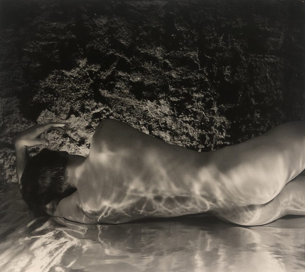 This is a warm-toned black-and-white photograph of a naked man lying with his back towards the camera with reflective material on the ground casting light on his body.