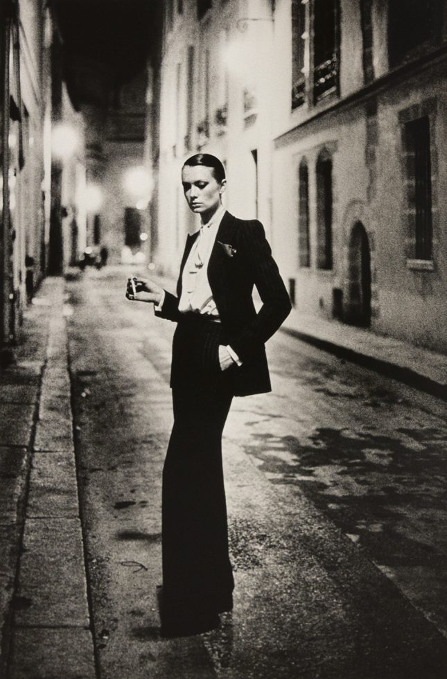 This is a warm-toned black-and-white photograph of a thin woman in a tuxedo with her hair slicked back standing in an urban street.