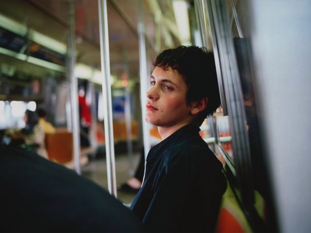 A young man sitting on the NYC subway leaning his back looking at a person to his left