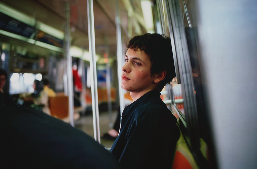 A young man sitting on the NYC subway leaning his back looking at a person to his left