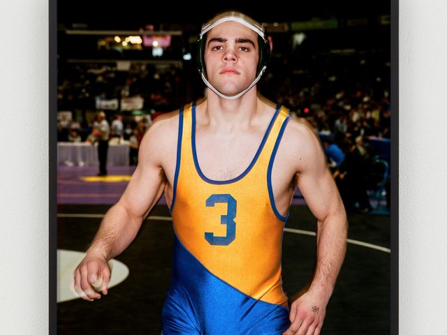 This is a photographic portrait of a young male wrestler wearing a blue and orange singlet.