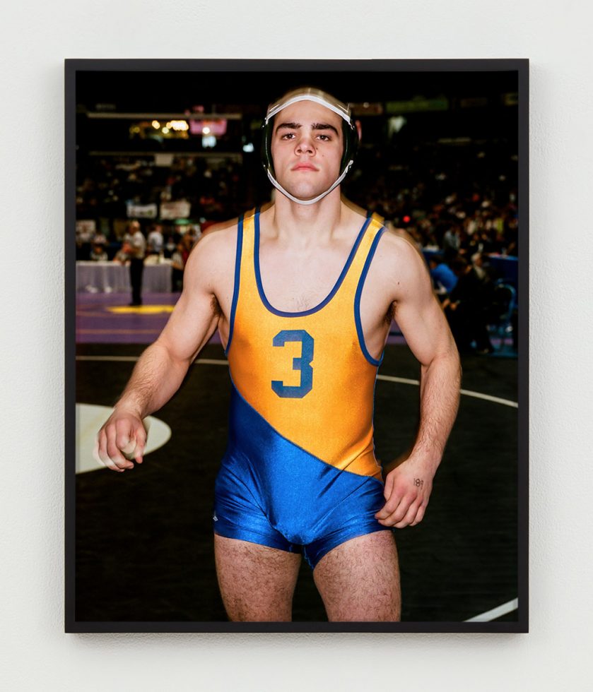 This is a photographic portrait of a young male wrestler wearing a blue and orange singlet.