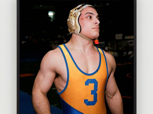 This is a photographic portrait of a young male wrestler with a blue and orange uniform and gold head gear.