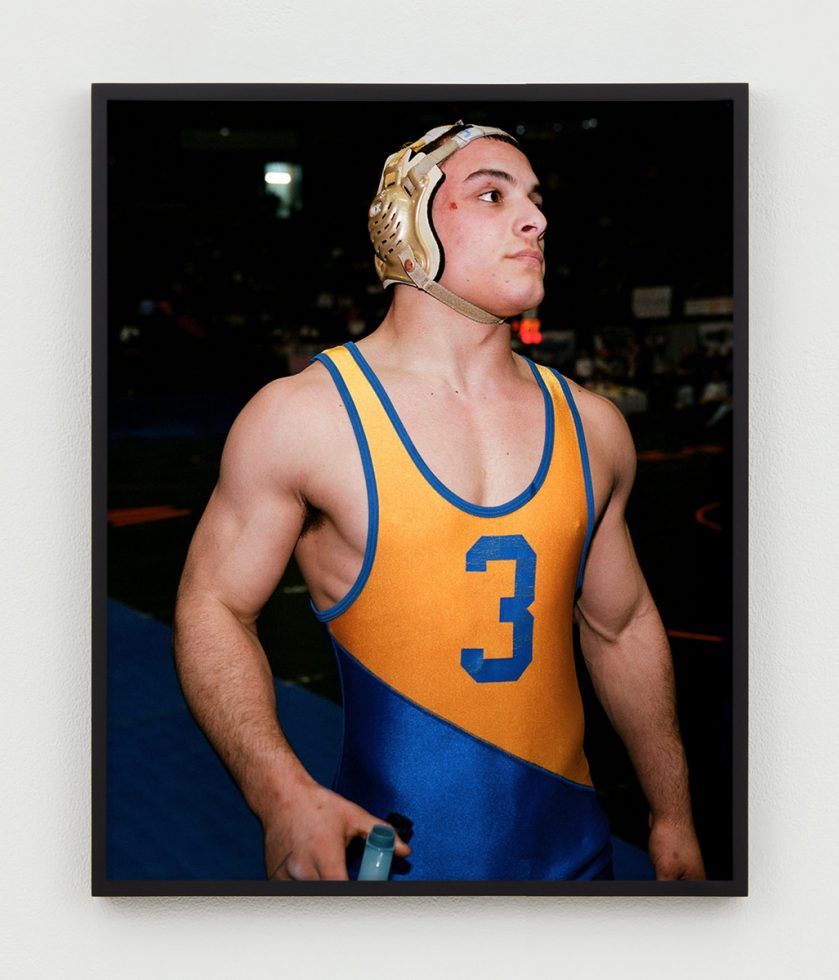 This is a photographic portrait of a young male wrestler with a blue and orange uniform and gold head gear.