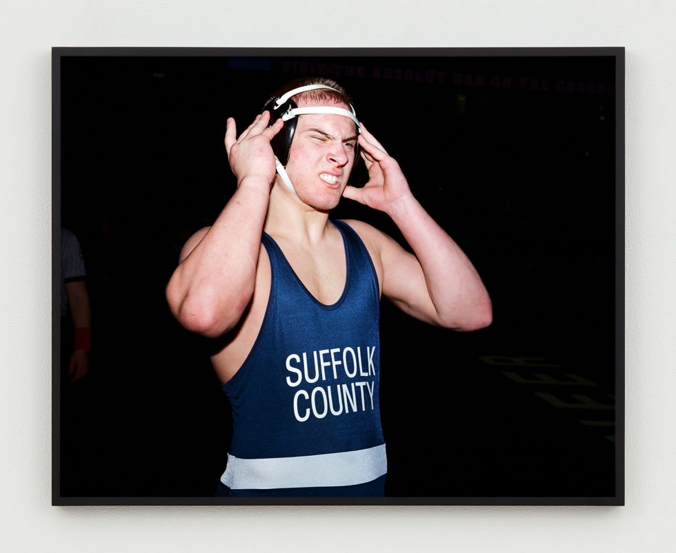 This is a photograph of a wrestler with a slightly bloody nose, wearing a “Suffolk County” singlet adjusting his head gear.