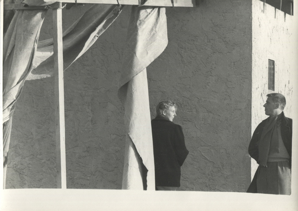 This is a black-and-white photograph of a woman and a man looking at each other behind some hanging fabric and in front of a building.