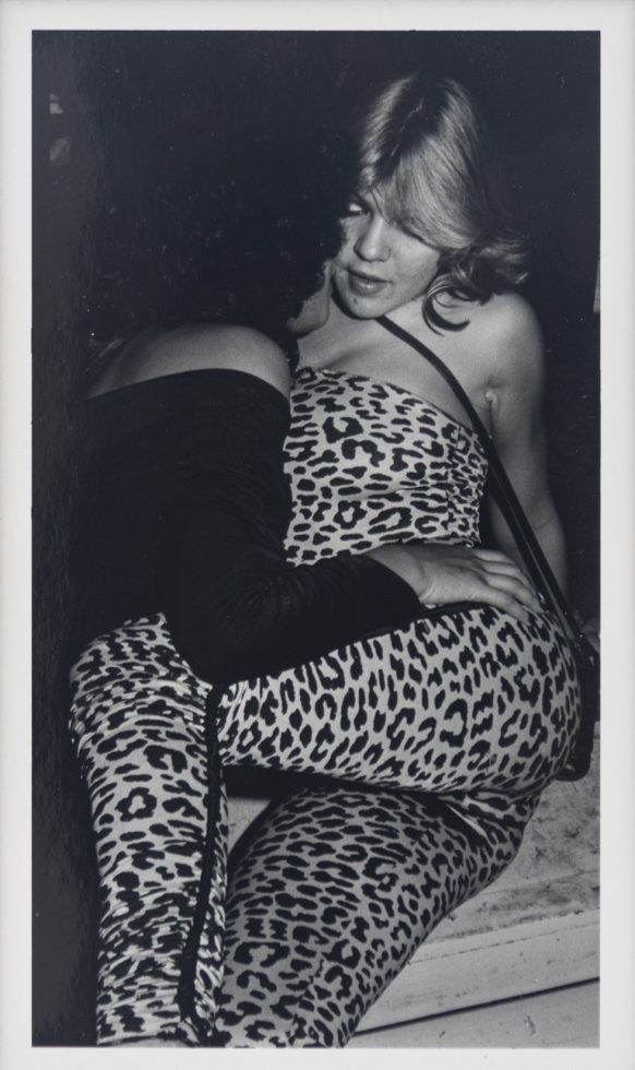 This is a black-and-white photograph of a blonde woman in an animal print bodysuit being embraced by another person.