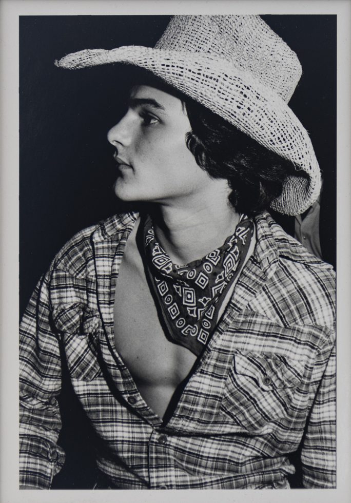 This is a black-and-white photograph of a young man in a plaid shirt, neckerchief, and cowboy hat.