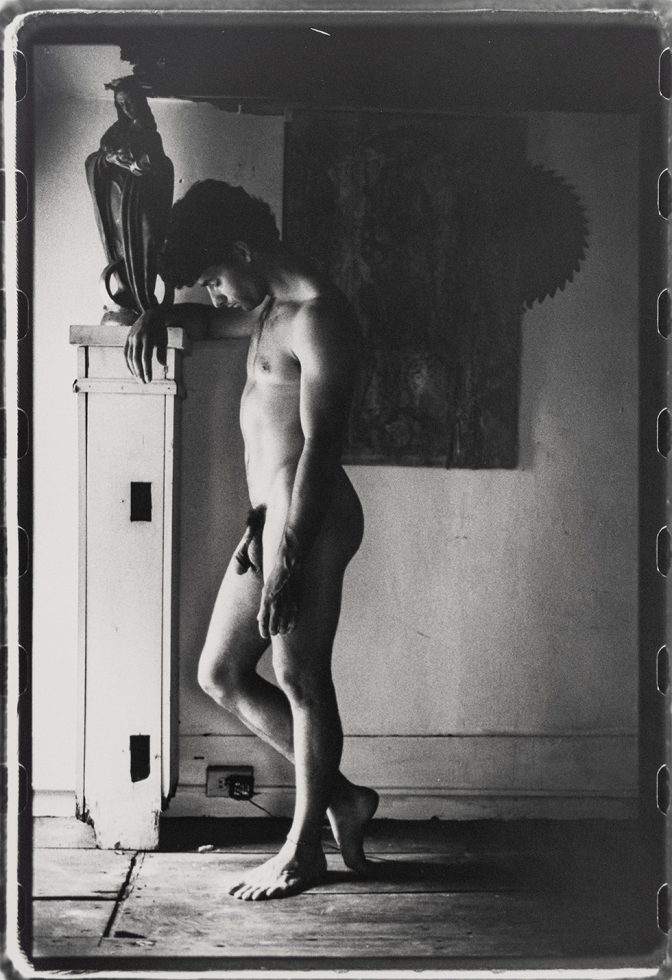 This is a black-and-white photograph of a naked man in profile leaning against a statue of the Virgin Mary.