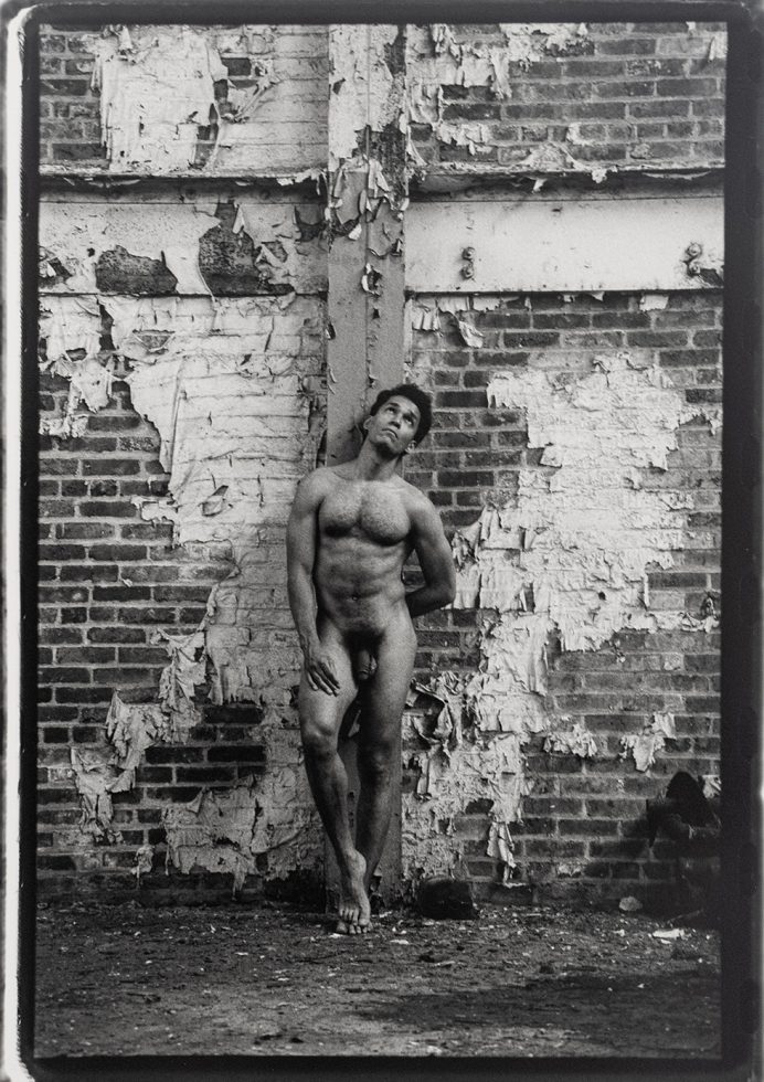 This is a black-and-white photograph of a naked man against a peeling bring wall.